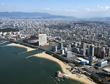 写真：上空から、福岡市内を眺めた様子。海沿いには、防波堤や砂浜、河川もありドームもある。高層マンションも海沿いに
多くある。たくさんの街並みが広がっている。